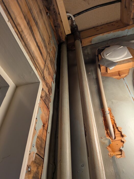 Rör i vägg med synliga skador och tecken på fukt. Väggpanel delvis borttagen, och en liten läcka syns vid ett äldre värmerör i ett badrum under renovering.