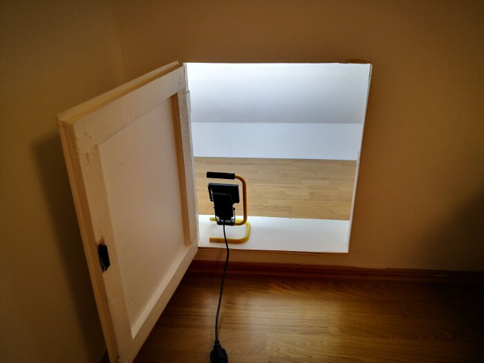 Lucka på väggen in till renoverat utrymme med parkettgolv och lampa som lyser upp rummet.