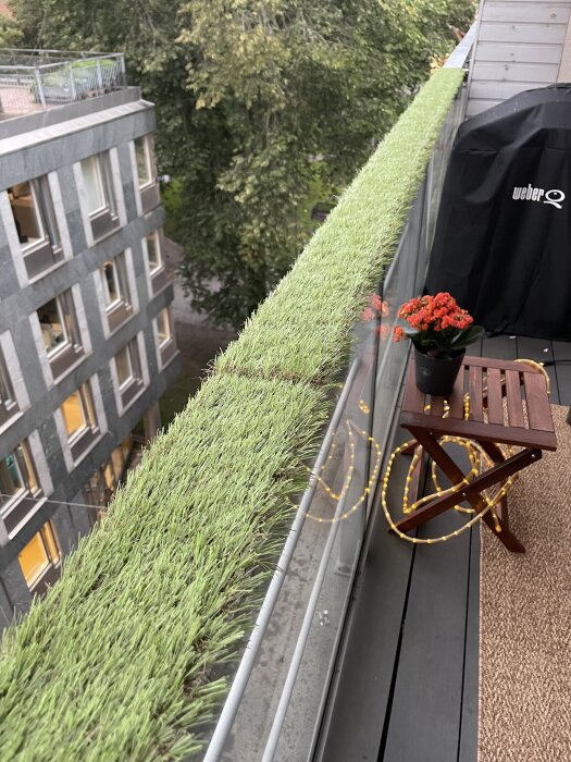 Konstgräs monterat på balkongräcke, en grill täckt med skydd, träbord med blommor ovanpå och utsikt mot en byggnad med träd i bakgrunden.