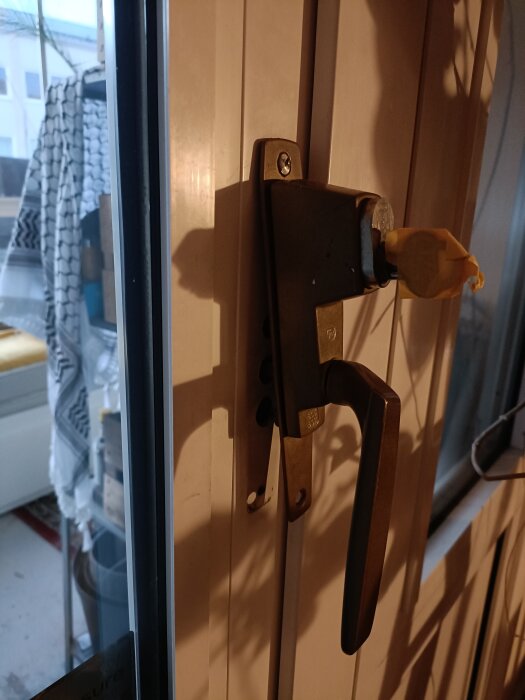 Handtag på inglasad balkongdörr med tejp och en envägsskruv i beslag. Bakgrund visar inredning på balkongen.