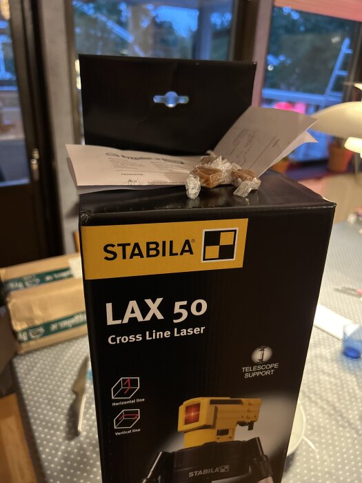 Förpackning av Stabila LAX 50 korslaser med några inslagna kolor ovanpå samt ett papper i bakgrunden.