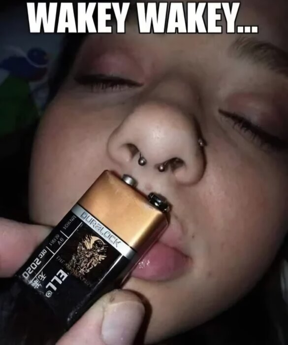 En 9V-batteri hålls mot läpparna på en person med ansiktspiercing, ögonen är stängda. Texten "Wakey Wakey..." visas ovanför.