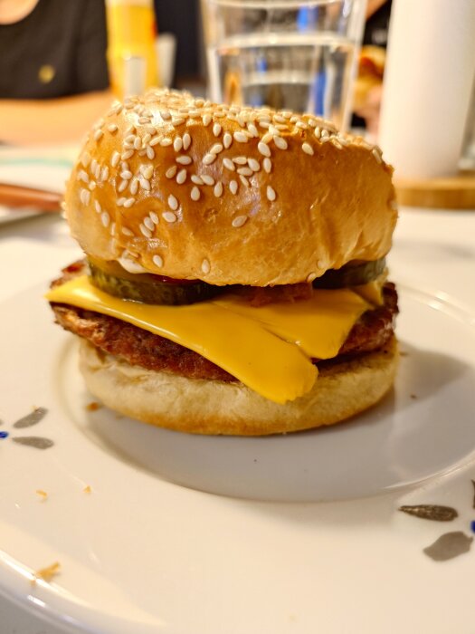 Hamburgare med ost och pickles serverad på en tallrik. Bakgrunden visar ett glas vatten och suddig bakgrundsmiljö.