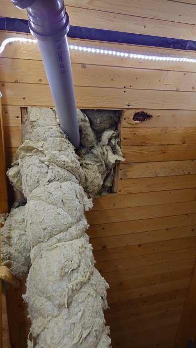 Bild av ett isolerat rör som går in i taket, omgiven av träpaneler och isolering. Rörsystem för avluftning i husets vind.