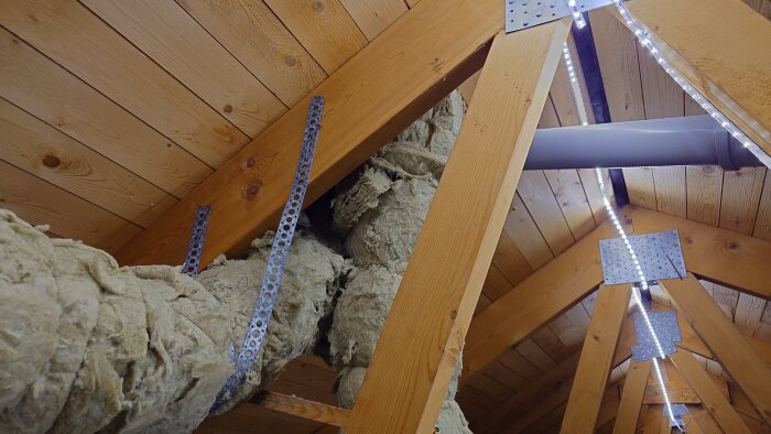 Isolerade ventilationsrör och träreglar på en vind med belysning och synliga skruvbeslag.