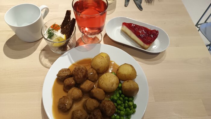 Svensk måltid med köttbullar, potatis, ärtor och sås på en tallrik, dessert i form av tårta, drink och förrätt på ett bord.
