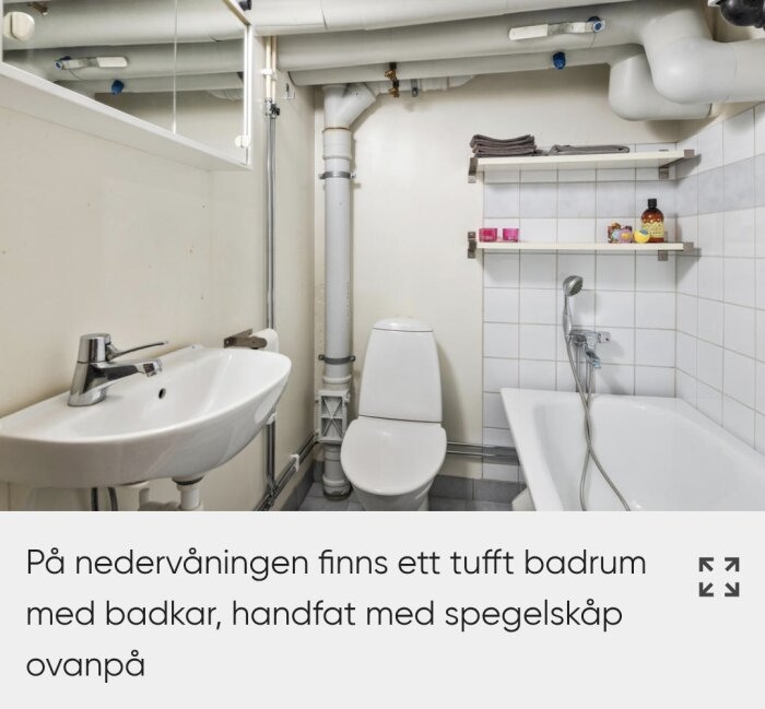 Badrum med badkar, handfat, spegelskåp och toalett, industristil med synliga rör och lagerhyllor med handdukar och badprodukter.