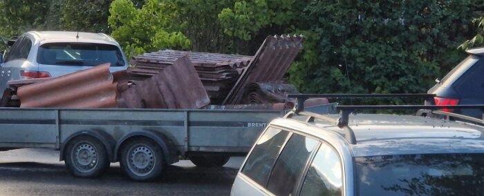 Släpvagn lastad med takplåt från ett husbyte, körs av bil på väg utan extra säkerheter som spännband. Grönska i bakgrunden.