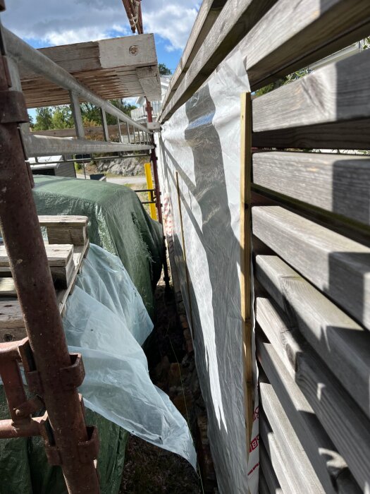 Träplank med genomskinlig plast, stött av stege och täckta ytor, visar ett byggprojekt eller staket med ändrat utseende.