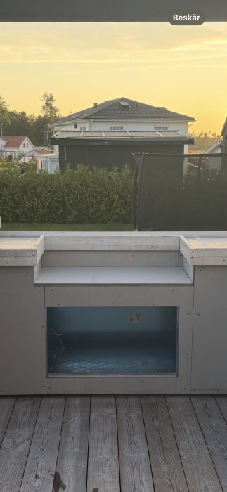 Utekök med stålreglar klädd med fibercementskivor, platsgjuten bänkyta, omgiven av trädgård med hus i bakgrunden vid solnedgång.