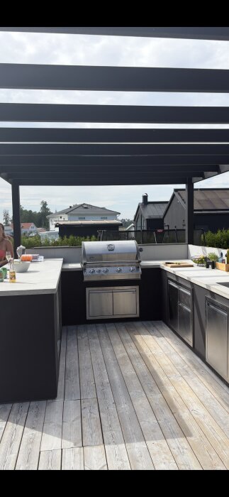 Utekök med stålgrill, gjuten betongbänk och takpergola, i en modern miljö med grönska och trähus i bakgrunden.