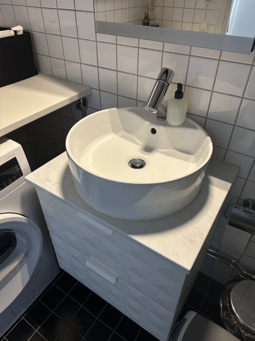 Modern vit badrumskommod med rund handfat, vitkaklade väggar och en svart tvättmaskin i bakgrunden.
