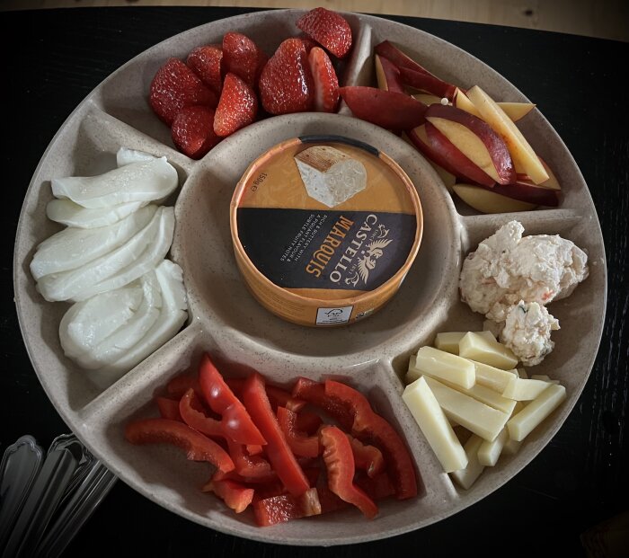 Ostbricka med olika ostar, jordgubbar, äppelklyftor, mozzarella, röd paprika och krämiga röror i sektionerade fack på ett rundfat.