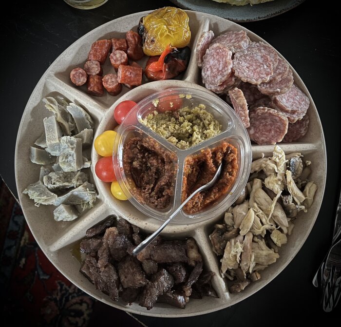 En tallrik med charkuterier, ostar, inlagda grönsaker och tre olika dippsåser, serverad på ett mörkt bord.