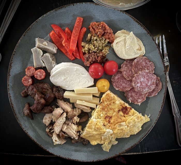 En tallrik med charkuterier, olika ostar, grönsaker som paprikor och tomater, samt en bit omelett.