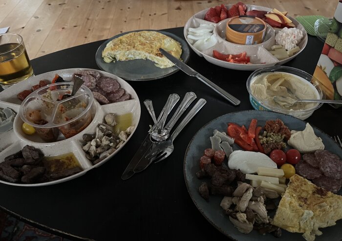 En måltid med olika maträtter, inklusive omelett, charkuterier, ost och grönsaker på flera tallrikar, samt ett glas dryck och en skål med hummus.