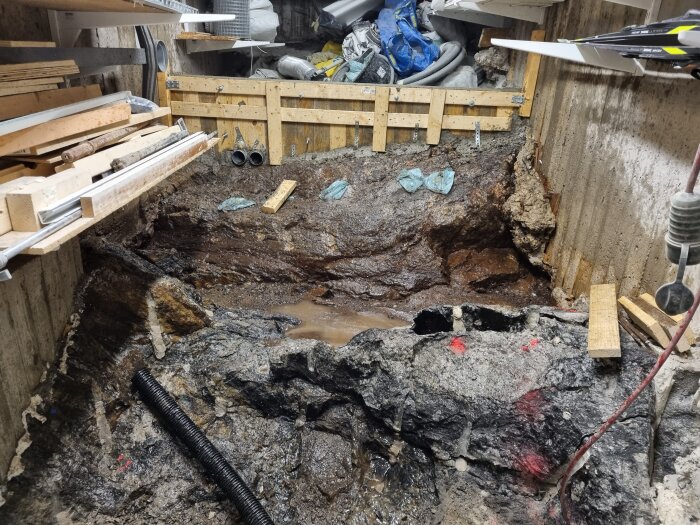 Byggarbetsplats med grävd grop i betonggolv. Omkring ligger träreglar och verktyg. Plaströr och diverse byggmaterial är synliga i bakgrunden.