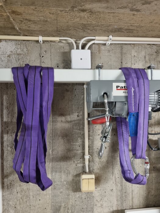 Lila remmar hänger över en metallbalk framför en betongvägg, nätverkskablar och en givarkabel synliga.