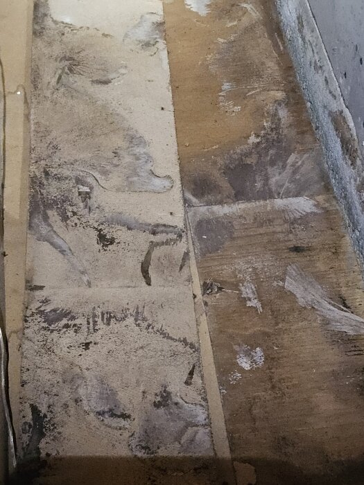 Slitet golv med fläckar och märken, troligen av murbruk eller lim, på en yta av träskivor nära en vägg.