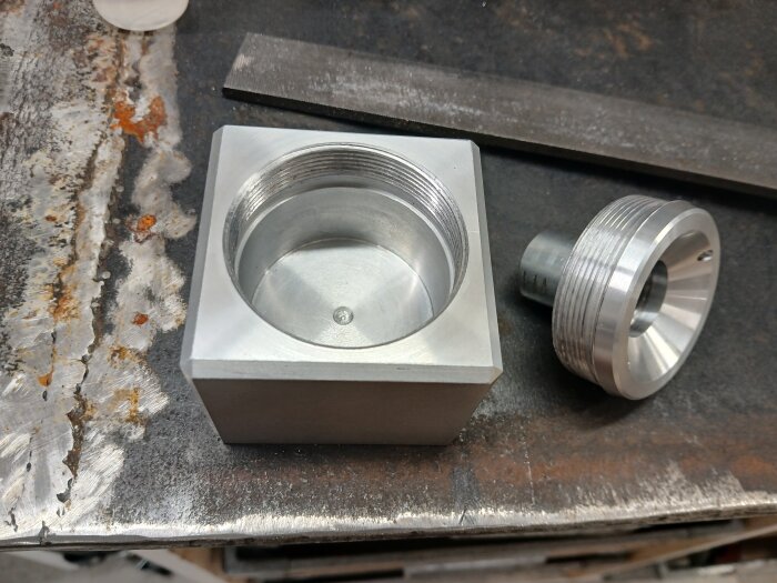 En metallcylinder med gängor bredvid en fyrkantig metallbehållare, båda verktyg för att hålla skärolja, placerade på en arbetsbänk.