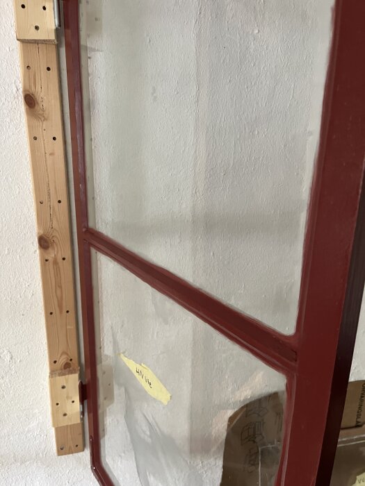 Ytterbåge med rödmålad trälist och kittad ruta, noterad med tejp. Vilar mot vit vägg och träkonstruktion i bakgrunden.