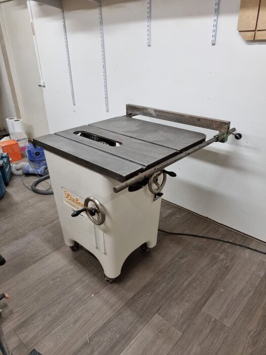 Bordssåg i vitt snickeri med metallbord, stående på trägolv. Bakom bordet syns väggmonterade hyllskenor och en sladd på golvet.