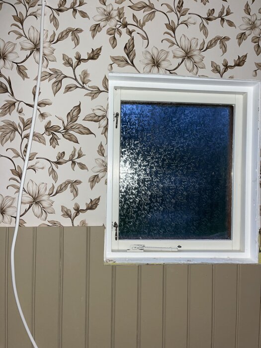 Vägg med blommig tapet och pärlspont vid ett fönster med vit karm i ett badrum.