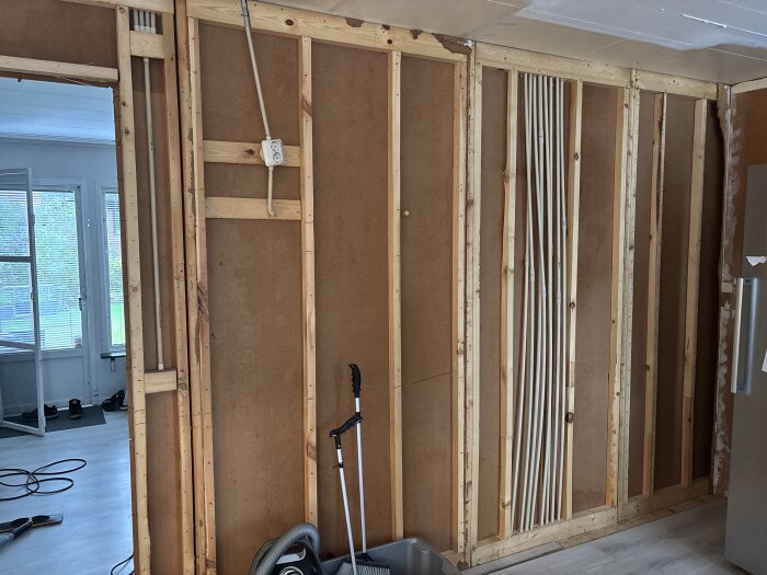 Vägg under renovering med synliga träreglar och rördragning, eluttag på väggen. Städskaft och dammsugare står intill.
