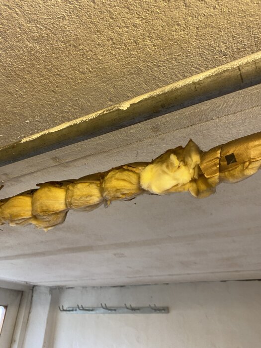 Gult isoleringsmaterial runt ett rör i taket i en källare. Rören verkar vara gamla och slitna, vilket väcker oro för asbestförekomst.