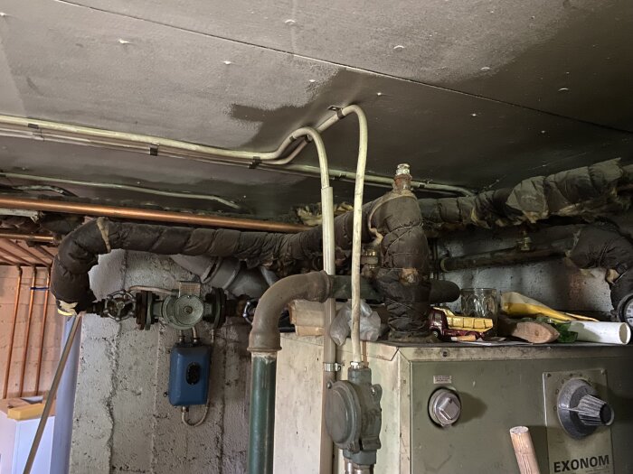 Gammal oljepanna i en källare omgiven av rör täckta med slitna isoleringsmaterial, potentiellt innehållande asbest.