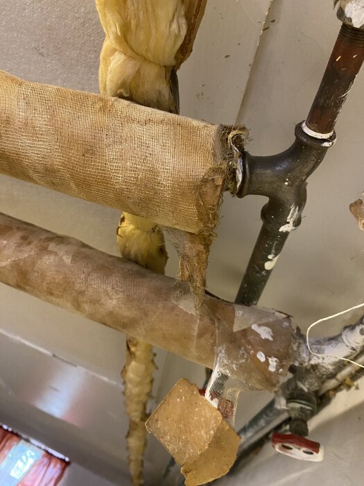 Rör i källartak med skadad isolering, potentiellt innehållande asbest, synliga kring en gammal oljepanna i ett hus från 1930-talet med 1970-talsutbyggnad.