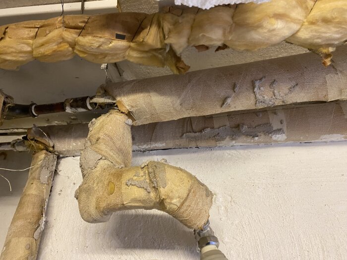 Skadade isolerade rör i källartaket med utsliten täckning och delvis blottade ytor, potentiellt risk för asbest.
