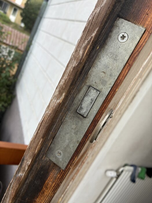 Närbild av en trädörrs kant med ett metallplattlås utan trycke, synlig utanför hus med tegelvägg i bakgrunden.