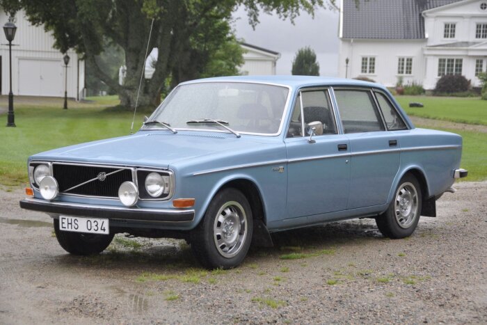 Blå Volvo 144 årsmodell 1971 parkerad framför ett vitt hus i grönskande miljö.
