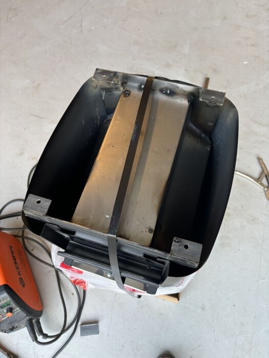 Svetsade vinkeljärn som fästpunkter på ett aggregat, med en oranget Kemppi-svetsaggregat och sladdar synliga på golvet bredvid.