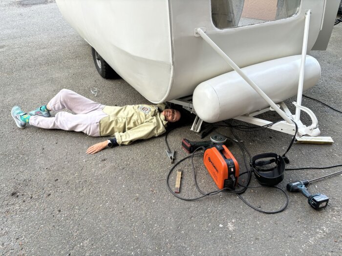 En person ligger under en husvagn, omgiven av verktyg som en cirkelsåg och svets, förbereder infästning av ett aggregat.