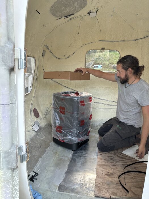 Person i husvagn installerar ett Harvia M3 bastuaggregat. Aggregatet är inplastat och placerat på golvet.