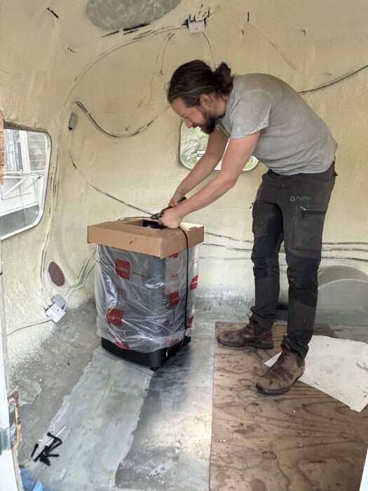 Man installerar ett Harvia M3-aggregat i husvagn med plywoodgolv. Aggregatet är delvis i förpackning, redo för montering.