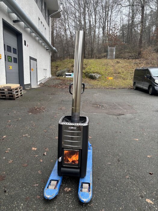 Vedeldat bastuaggregat från Harvia står på en blå palldragare utomhus. Skorstenen installeras och en eld brinner inuti aggregatet.