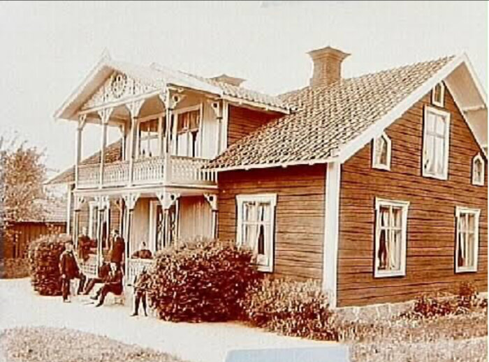 Stor träbyggnad med snickeridetaljer och balkong, omgiven av buskar. Flera personer står och sitter framför huset.