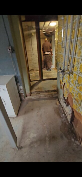 Källargolv med nivåskillnader, rör på vägg med blommig tapet, person i korridor, tvättmaskin till vänster.