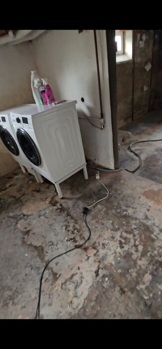 Tvättmaskin och torktumlare i ett slitet källarutrymme med ojämnt golv och kablar liggande på marken. Rör synliga på väggen.