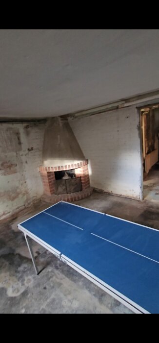 Öppen spis i källare med tegelväggar och ett pingisbord i förgrunden. Ojämnt betonggolv syns.