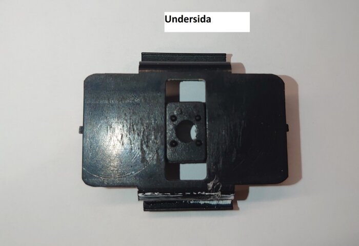 Svart plastclip med monteringshål, märkt "Undersida", används för att fästa aluminiumprofiler på balkong; letar efter ersättningsclips.