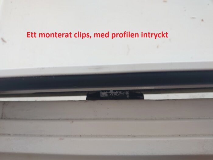 Ett monterat clips med en aluminiumprofil inskjuten, används för att fästa fodren vid en karm, svår att ta bort utan att clipsen går sönder.