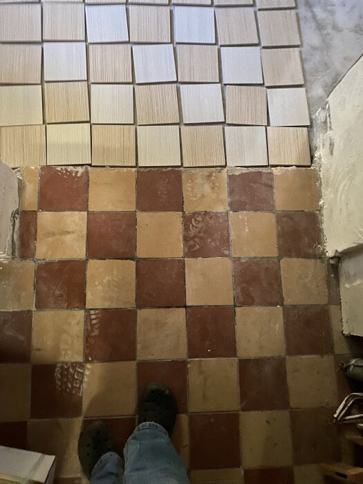 Rutigt golv med rödbruna och gula plattor, jämförs med ljusare, oregelbundet placerade plattor. En persons fötter syns vid kanten.