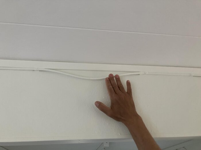 En hand vidrör en utanpåliggande vit elkabel som är fäst mot ett vitt tak, men hänger slakt i mitten.