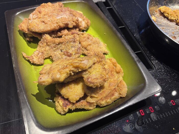 Fläskschnitzel på grön ugnsform med en svart stekpanna bredvid.