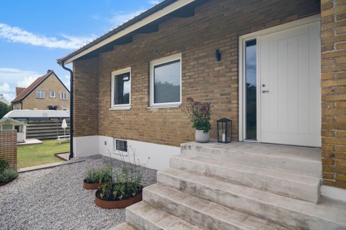 Vit ytterdörr med glaspanel syns på ett brunt tegelhus. Framför finns en betongtrappa med blomkruka och lykta, intill en grusgång.