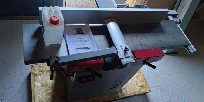 Hammer A3 snickerimaskin på golvet med manual och plastdunk ovanpå, omgiven av verkstadsdetaljer.